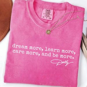 Könnte beinhalten: Ein leuchtend rosa T-Shirt mit kurzen Ärmeln und dem weißen Aufdruck: dream more, learn more, care more, and be more. Das T-Shirt zeigt unter dem Text eine Unterschrift im Stil des Namens Dolly sowie ein Markenetikett von Comfort Colors.