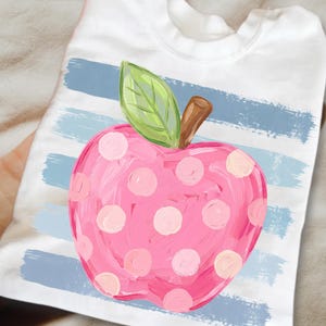 Peut inclure: Un t-shirt blanc avec un motif de pomme rose à pois rose clair, une feuille verte et une tige brune. La pomme est superposée sur un fond de rayures horizontales texturées en forme de coups de pinceau bleu clair.