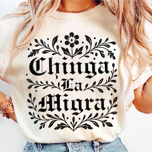 May include: Cream-colored t-shirt with the phrase "Chinga La Migra" in bold, black gothic lettering, framed by floral and leaf designs. The design includes a central flower and symmetrical leaf patterns.