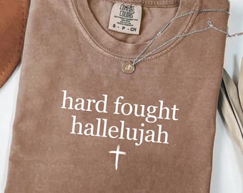 May include: A brown Comfort Colors t-shirt with the words "hard fought hallelujah" and a cross printed in white. A silver necklace with a pendant rests on the shirt.