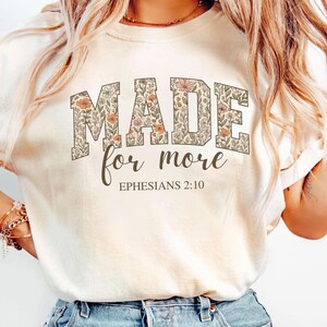 Op de afbeelding: Een wit T-shirt met een bloemenprint met de tekst "MADE for more" en "EPHESIANS 2:10" in bruine letters.