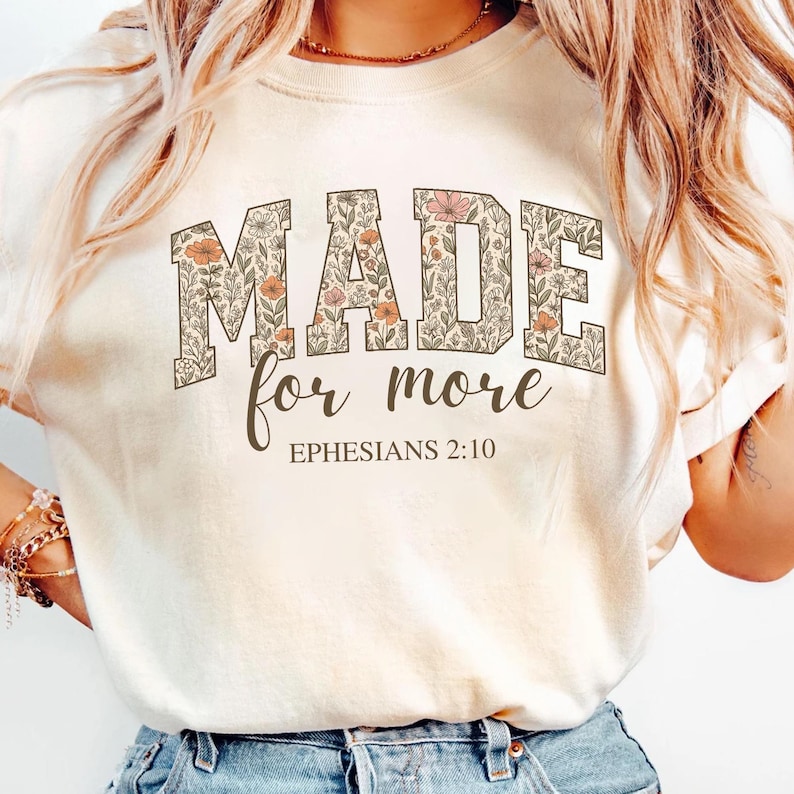 Op de afbeelding: Een wit T-shirt met een bloemenprint met de tekst "MADE for more" en "EPHESIANS 2:10" in bruine letters.