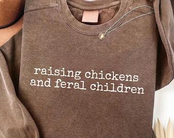 May include: A brown sweatshirt with the text "raising chickens and feral children" in white. The sweatshirt has a crew neck and a small tag near the neckline. A silver necklace with a small pendant is draped across the front.