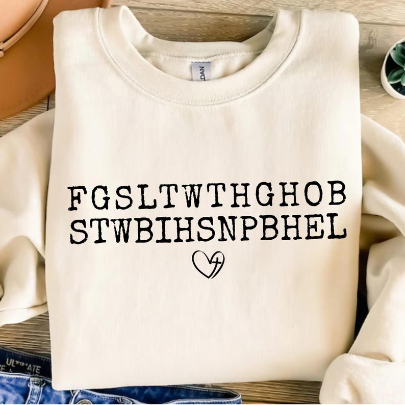 May include: Cream-colored sweatshirt with black text and a heart with a cross graphic. The text is arranged in two rows of capital letters. The sweatshirt is folded and placed on a surface with a hat and a plant in the background.