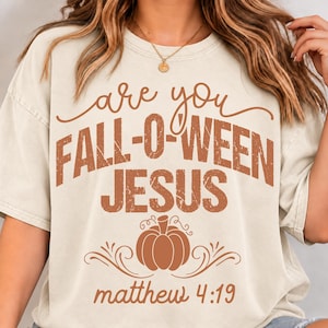 May include: A cream-colored short-sleeve t-shirt featuring brown text that reads are you FALL-O-WEEN JESUS and matthew 4:19. The design includes a central pumpkin graphic with decorative swirls above and below the pumpkin illustration.
