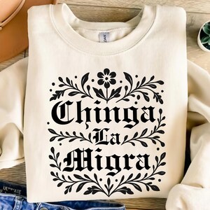 May include: Cream-colored sweatshirt with the phrase "Chinga La Migra" in black gothic lettering, framed by a floral and leaf design. The sweatshirt is a casual, long-sleeved garment.