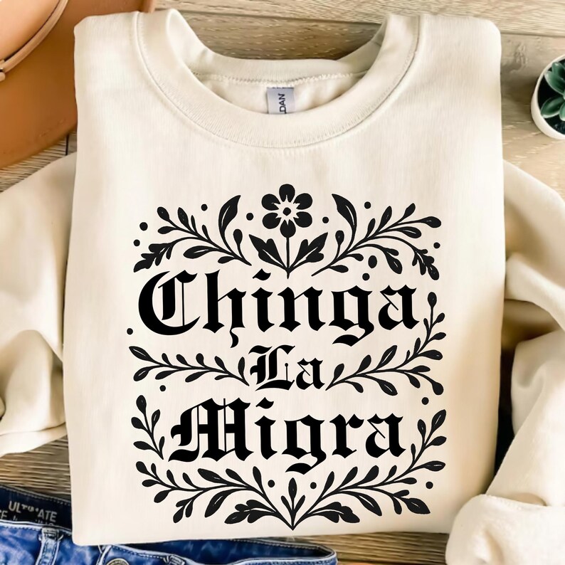 May include: Cream-colored sweatshirt with the phrase "Chinga La Migra" in black gothic lettering, framed by a floral and leaf design. The sweatshirt is a casual, long-sleeved garment.