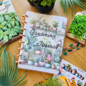 May include: A gardening journal with a watercolor illustration of potted plants on the cover. The journal has a gold spiral binding and the words "Gardening Journal" are printed in black on the cover.