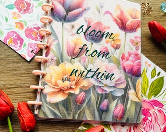 May include: A pink and white floral design planner with a clear plastic cover. The planner has a pink and white floral design with the words "bloom from within" in green text.