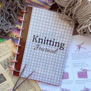 Peut inclure: Un carnet "Knitting Journal" avec une tranche marron et des anneaux dorés, ouvert sur une page avec des instructions de tricot. De la laine grise et des aiguilles à tricoter sont visibles, ainsi que d'autres fournitures.