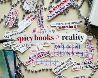 May include: A collection of colorful stickers with text about books and reading. The stickers are arranged on a white surface with a silver beaded garland. Some of the text on the stickers includes "Spicy books > reality", "Books are proof humans can do magic", and "I'm here for it".