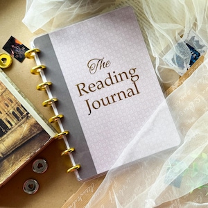 Może przedstawiać: Dziennik czytelnika z oprawą dyskową, jasnolawendową okładką z subtelnym tkanym wzorem i złotym napisem The Reading Journal. Notatnik posiada ciemnoszary pasek na grzbiecie oraz błyszczące, złote metalowe dyski wzdłuż lewej krawędzi.
