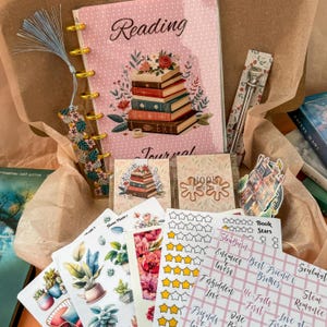 Op de afbeelding: Een roze en gouden leesdagboek met een illustratie van een stapel boeken, een bladwijzer, een pen en verschillende stickers. De stickers omvatten een huis, een boekbeoordelingstabel, een bloemenontwerp en een plantenvel met stickers.