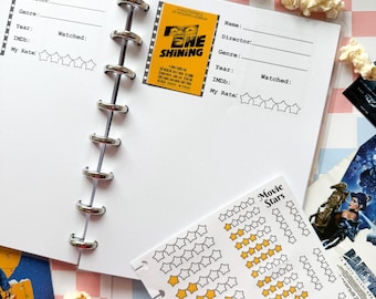 May include: A white planner with a black and white movie ticket sticker for the movie "The Shining" on the right page. The left page has a section for movie details, including name, director, genre, year, IMDb rating, and a section to rate the movie with five stars. The planner has a silver ring binding. The planner is on a pink and white checkered background with popcorn.