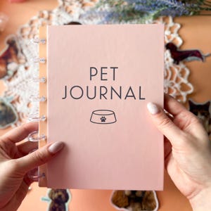 May include: A pink Pet Journal with the words "PET JOURNAL" and a paw print bowl graphic. The journal is held open, revealing its spiral binding. Decorative elements and pet stickers are in the background.