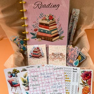 Könnte beinhalten: Ein rosa Planer mit einer goldenen Spirale und einem Stapel Bücher mit floralen Akzenten. Der Planer hat das Wort "Lesen" auf der Vorderseite. Das Bild enthält auch eine Vielzahl von Aufklebern, darunter ein Haus, einen Stapel Bücher und ein Blumenmuster.