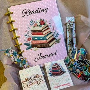 Op de afbeelding: Een roze leesdagboek met gouden ringen en een illustratie van een stapel boeken. Het dagboek is omringd door andere boekgerelateerde items, waaronder een bladwijzer, een pen en een sticker. De sticker zegt "Cozy Nights".