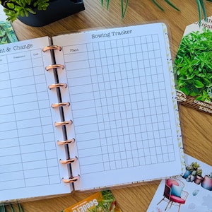 May include: A white planner with a gridded page for tracking sowing dates. The page is labeled "Sowing Tracker" and has a column for "Plant". The planner has a rose gold binding.