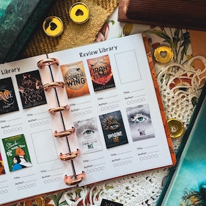 May include: A journal with a rose gold binding, open to a page titled "Review Library." The page features book covers with titles like "Fourth Wing" and "Iron Flame." Gold heart-shaped paper clips and decorative elements surround the journal.