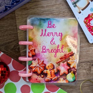 Op de afbeelding: Een roze en transparante plastic map met een aquarelontwerp van peperkoekjes en de tekst "Be Merry & Bright".