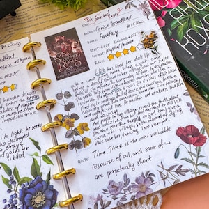 Może przedstawiać: Strona w notatniku na dyskach z odręcznymi notatkami z recenzji książki Six Scorched Roses autorstwa Carissy Broadbent. Strona zawiera wydrukowany obraz okładki książki, ocenę czterech gwiazdek, podsumowanie i cytat, ozdobione naklejkami kwiatowymi.