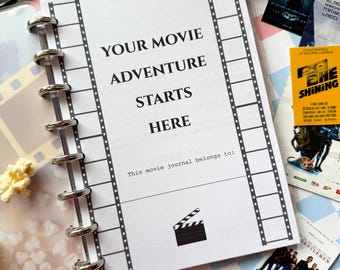 May include: A white journal with black film strip borders and the text "Your Movie Adventure Starts Here". The journal has a silver spiral binding and a black and white illustration of a clapperboard.