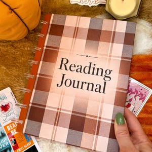May include: A disc-bound reading journal with a brown and cream plaid cover. The front cover features the text Reading Journal in a black serif font. The journal has a solid brown spine and clear plastic discs along the left side for binding.