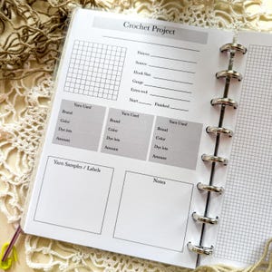 May include: Open notebook with a gray and white grid pattern and the words "Crochet Project" at the top. The notebook has sections for pattern details, yarn information, and notes. The notebook is open to a page with a grid and several sections for recording crochet project details.
