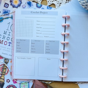 Op de afbeelding: Een roze en witte crochet project planner notebook met een raster voor patroondetails, gareninformatie en notities. Het notitieboek is open, met gelinieerde pagina's en een spiraalbinding. De cover heeft de tekst "Crochet Project".