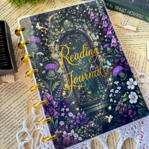 May include: A disc-bound reading journal featuring a dark, enchanted garden illustration with a central arched door, purple thistle flowers, and white blossoms. The cover displays the gold cursive text Reading Journal and has gold metal binding discs.