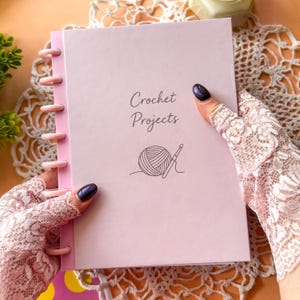 Op de afbeelding: Een lichtroze notitieboek met de titel "Crochet Projects" en een illustratie van een bol garen. Het notitieboek heeft een roze rug met ringband. Het notitieboek wordt vastgehouden door een persoon met kanten handschoenen.