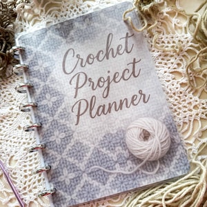 Op de afbeelding: Een Crochet Project Planner met een witte en grijze gebreide patroon cover. De planner is spiraalgebonden met zilveren ringen en bevat een bolletje witte wol. De tekst "Crochet Project Planner" staat op de cover.