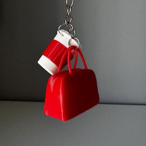 May include: A keychain featuring a miniature red handbag and a white and red coffee cup. The handbag has two small handles. The coffee cup has a red stripe around the middle. The keychain is silver.