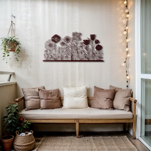 May include: A dark brown metal floral wall art piece featuring various flower silhouettes. The artwork is mounted on a light-colored wall above a wooden bench with pillows. A hanging plant and string lights add to the decor.