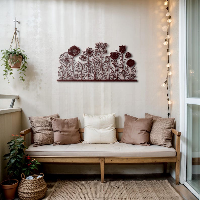 May include: A dark brown metal floral wall art piece featuring various flower silhouettes. The artwork is mounted on a light-colored wall above a wooden bench with pillows. A hanging plant and string lights add to the decor.