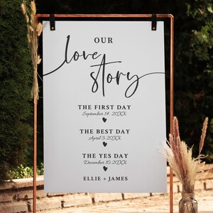 May include: A copper-colored metal wedding sign with a white background. The sign reads "Our Love Story" in elegant script, with dates and names below. Decorative dried flowers are in a glass vase to the right.