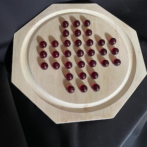 Peut inclure: Un plateau de jeu octogonal en bois clair doté d'une zone circulaire encastrée contenant une grille de trente-trois petites pièces de jeu sphériques, translucides et rouge foncé, disposées en croix pour un jeu de solitaire.
