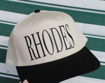 Może przedstawiać: Kremowa czapka bejsbolowa z czarnym daszkiem i napisem "RHODES" wydrukowanym dużymi, czarnymi literami. Czapka ma czarny guzik na górze. Tło to wzór w zielono-białe paski.