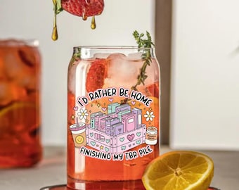 May include: A clear glass can-shaped tumbler filled with a red beverage, ice, and strawberries. The tumbler has a graphic that reads "I'D RATHER BE HOME FINISHING MY TBR PILE" with illustrations of books, flowers, and a coffee cup. A strawberry and lemon half are also visible.