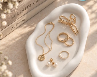 May include: A gold-toned jewelry set on a white organic-shaped tray, including a delicate chain necklace with a round pendant, a hair claw clip, two simple rings, and a pair of stud earrings featuring small white pearls.