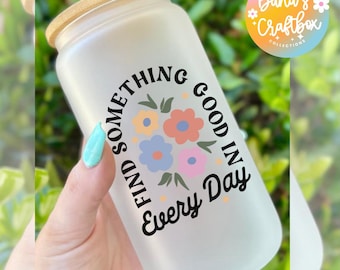 May include: Frosted glass tumbler with a bamboo lid. The tumbler features the text "Find Something Good in Every Day" with a floral design in the center. The glass is held by a hand with a light blue manicure.