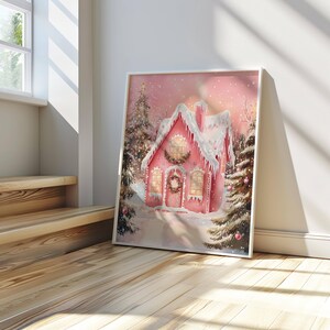 May include: A framed watercolor print of a pink gingerbread house covered in snow, with a wreath on the door and Christmas trees. The artwork is set against a soft pink background, evoking a winter holiday theme.