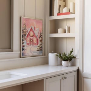 May include: A framed print of a pink gingerbread house in a snowy winter scene. The house has a wreath on the door and lights in the windows. The print is displayed on a white countertop next to a white ceramic container and a small potted plant.