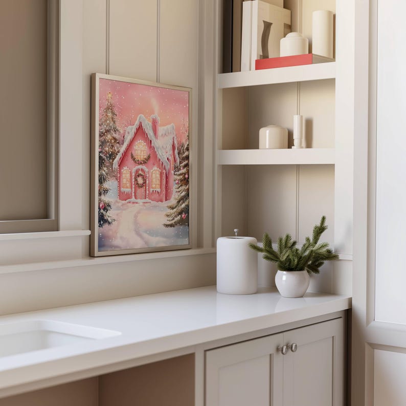 May include: A framed print of a pink gingerbread house in a snowy winter scene. The house has a wreath on the door and lights in the windows. The print is displayed on a white countertop next to a white ceramic container and a small potted plant.