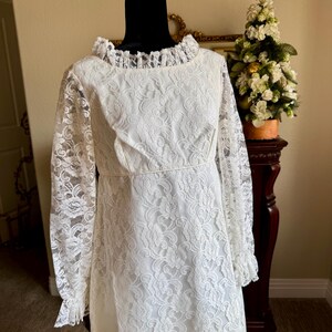 May include: A vintage white lace dress with long sleeves and a ruffled collar. The dress is displayed on a black mannequin and features a floral lace pattern. The dress has a fitted bodice and a flowing skirt, perfect for a special occasion.