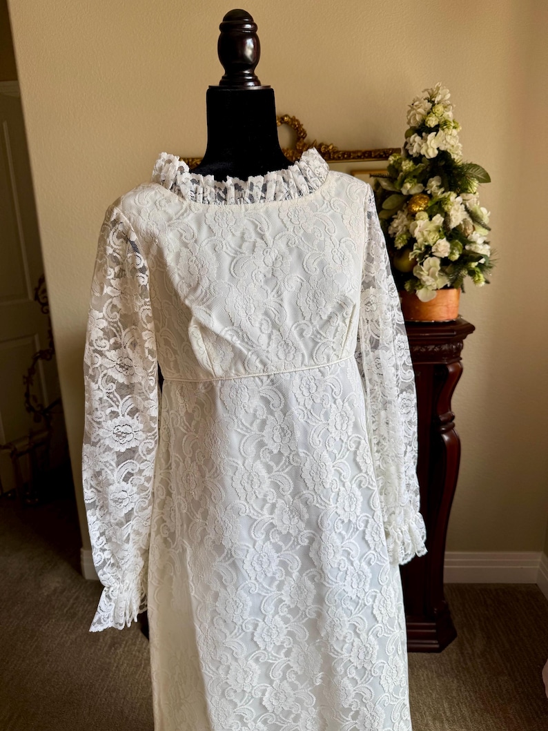 May include: A vintage white lace dress with long sleeves and a ruffled collar. The dress is displayed on a black mannequin and features a floral lace pattern. The dress has a fitted bodice and a flowing skirt, perfect for a special occasion.