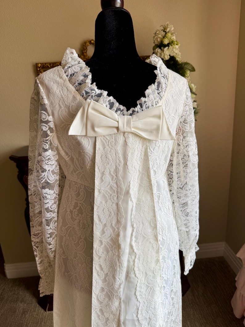 May include: Ivory lace dress with long sleeves and a large bow detail. The dress features a ruffled collar and a fitted bodice. The lace pattern is intricate, and the dress appears to be vintage or vintage-inspired. The dress is displayed on a black mannequin.