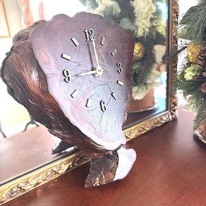 May include: A unique wooden clock with a natural, organic shape. The clock face is a dark, reddish-brown color with visible wood grain and cracks. Gold-colored hands and numerals mark the time. The clock is set on a wooden surface.