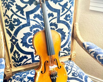 May include: A wooden violin with a brown varnish finish sits on a blue and white patterned chair. The violin has a black fingerboard and black tuning pegs.