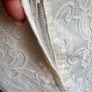 May include: Close-up of a white lace fabric with a zipper. The lace features a floral pattern. The zipper is white and partially open, revealing the fabric's texture and construction. The overall aesthetic is delicate and elegant.